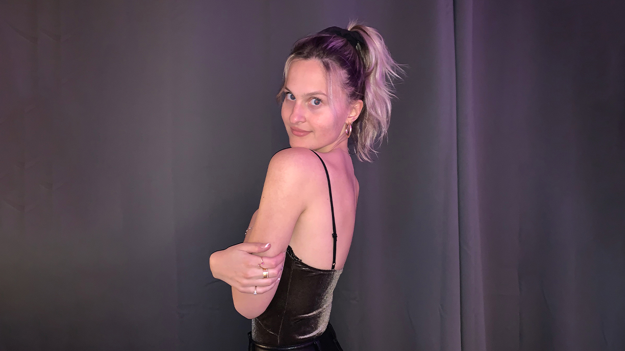 Photo of Danijela in black strappy tank facing away from the camera, looking back over her shoulder. Her blonde hair is in a ponytail and her arms are crossed over her chest. She's standing in front of a grey curtain. 