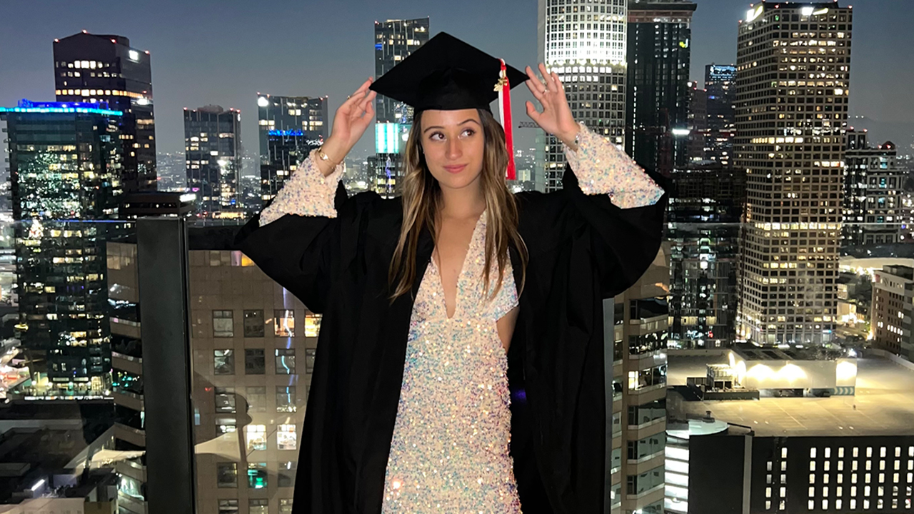 Photo of FIDM Graduate Nicole Rodopoulos in front of Downtown LA backdrop, wearing a sparkly dress and a graduation robe and cap. She's holding the cap on her head and eyeing the tassel hanging from it. 
