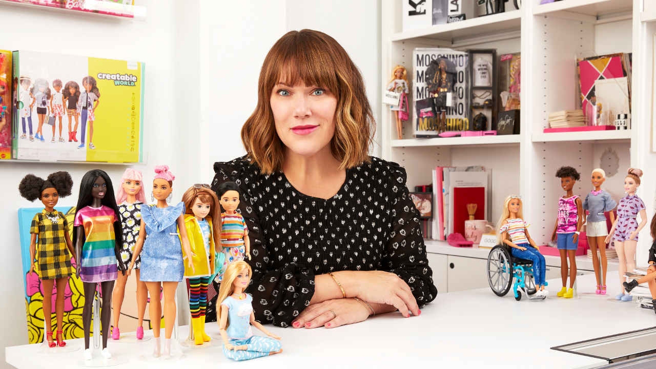 Kim Culmone sitting at her desk wearing a black long sleeve shirt, Barbie dolls on desk