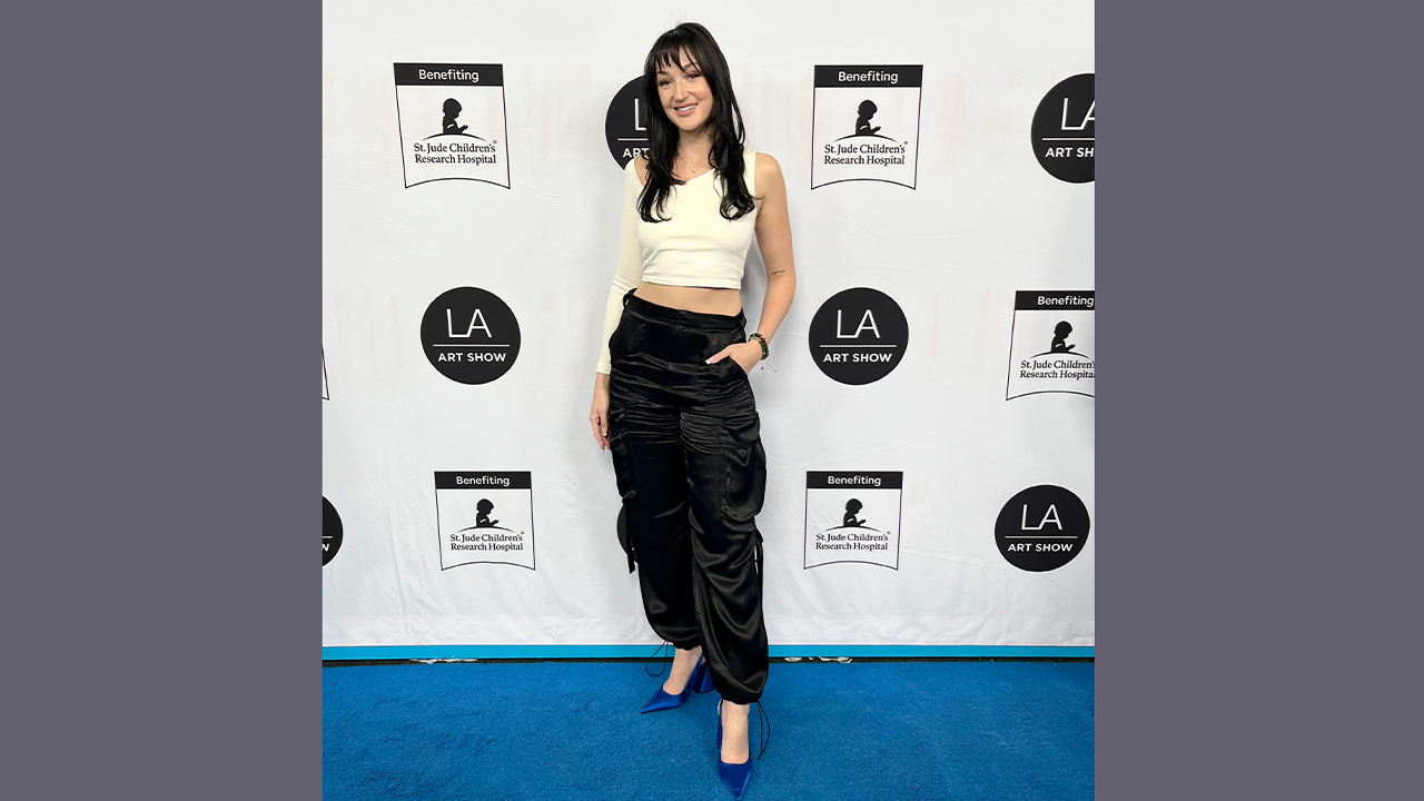 Jordyn is on the LA Art Show Step-and-repeat wearing black satin pants, blue heels, and a white cropped one-sleeve top.. 
