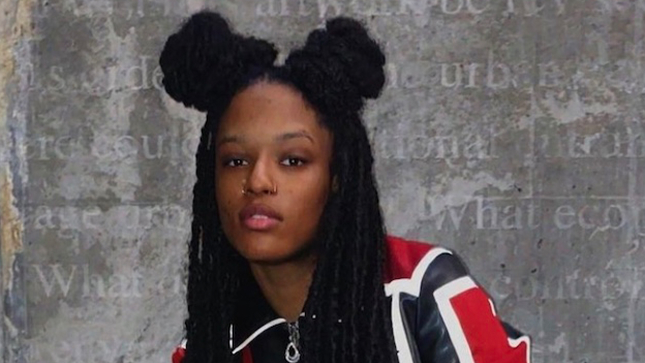 LRÉ gazes directly into the camera in front of a concrete wall with writing on it. Her hair is in braids, twisted up on either side of her head, and she's wearing a black, white, and red jacket.