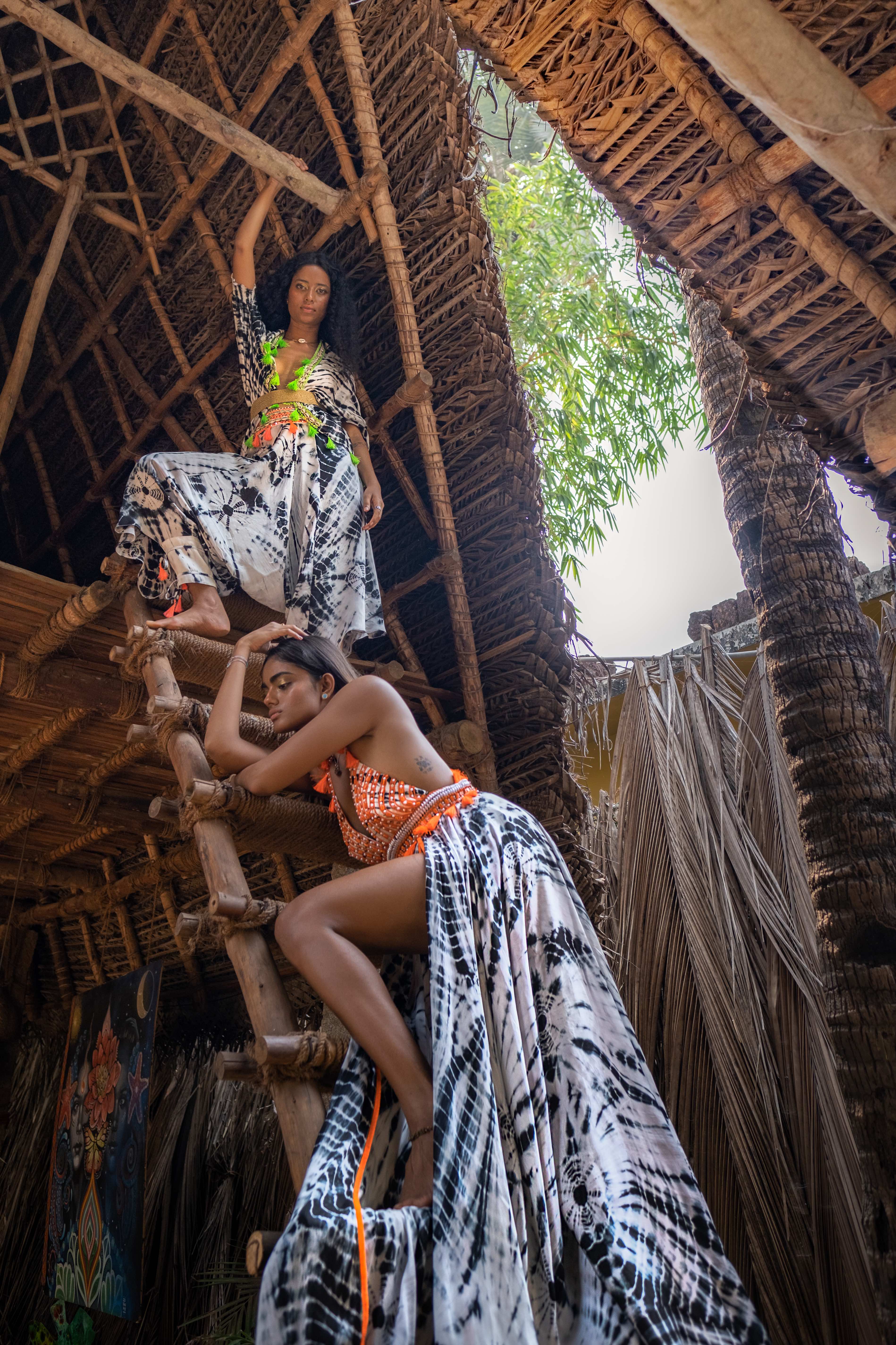 two models wear long white dresses with bright orange and yellow accents by Surily G standing on a ladder to a tree house outdoors