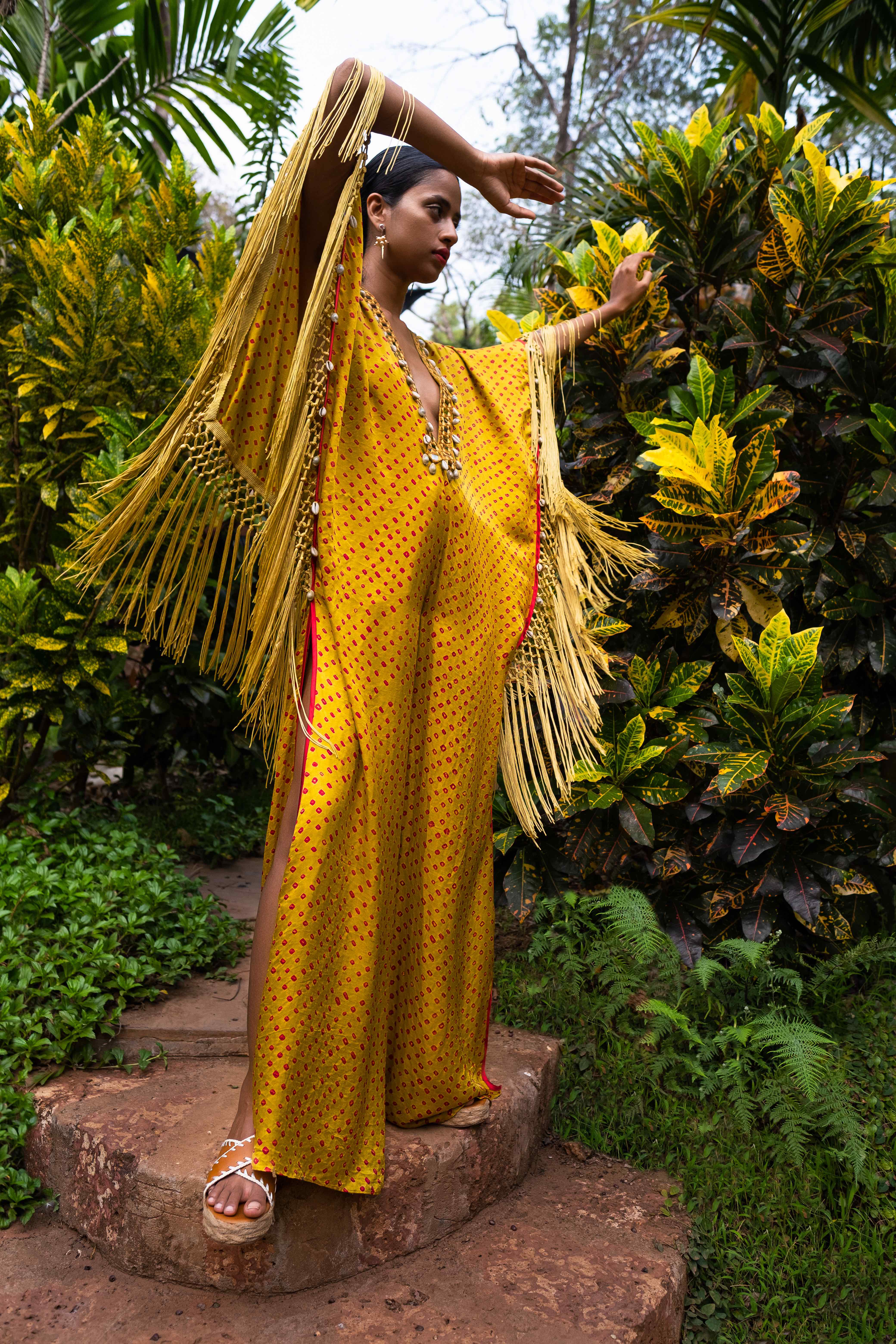 model wearing full-length yellow silk v-neck dress with fringe by Indian fashion designer Surily DP Goel standing outdoors in front of green lush plants