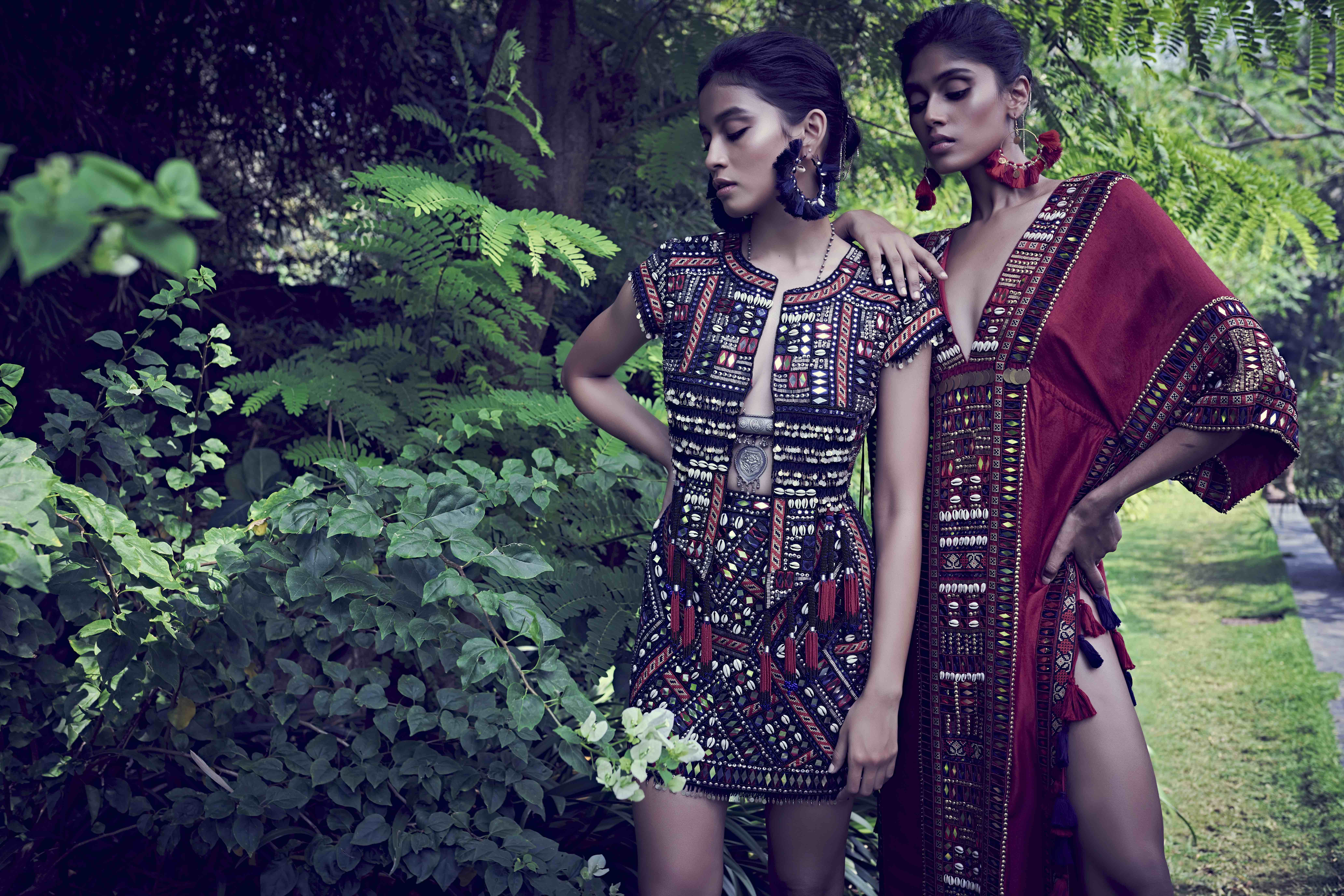 two models wear fashion designs by Surily G standing outside with lush green plants behind them as they look at the ground