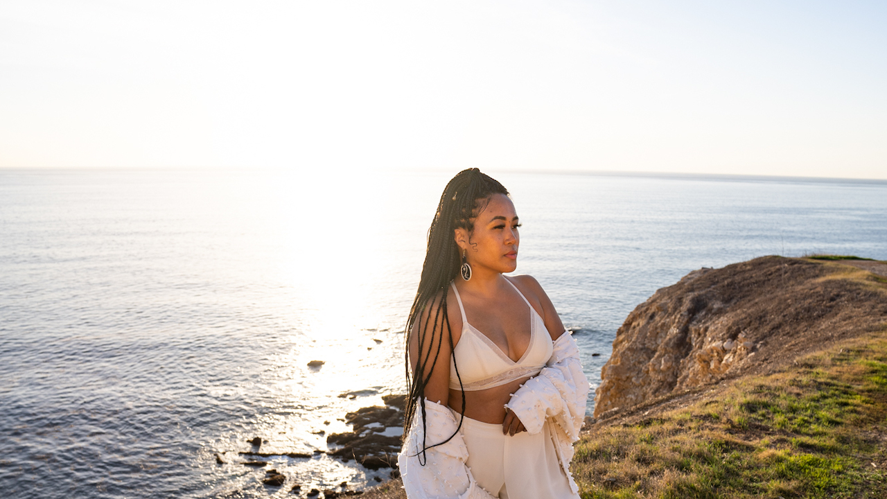 FIDM Grad Jen Benitez poses in a white bra top and white pants on a cliffside with the ocean behind her