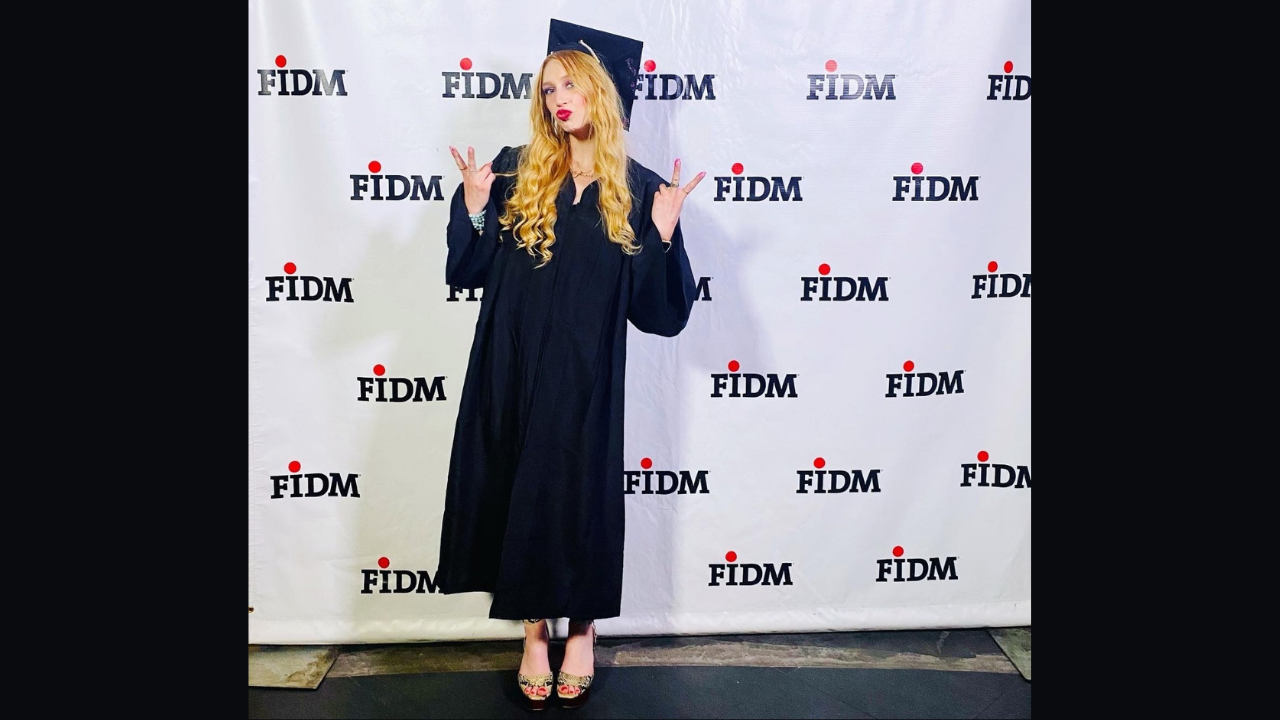 FIDM IMPD Grad Jessa Braunreiter stands in cap and gown in front of FIDM step and repeat