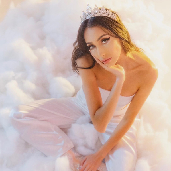 Photo of Ashlie looking up at camera wearing crown, white strapless top, and white sheer pants, sitting in a fluffy cloud