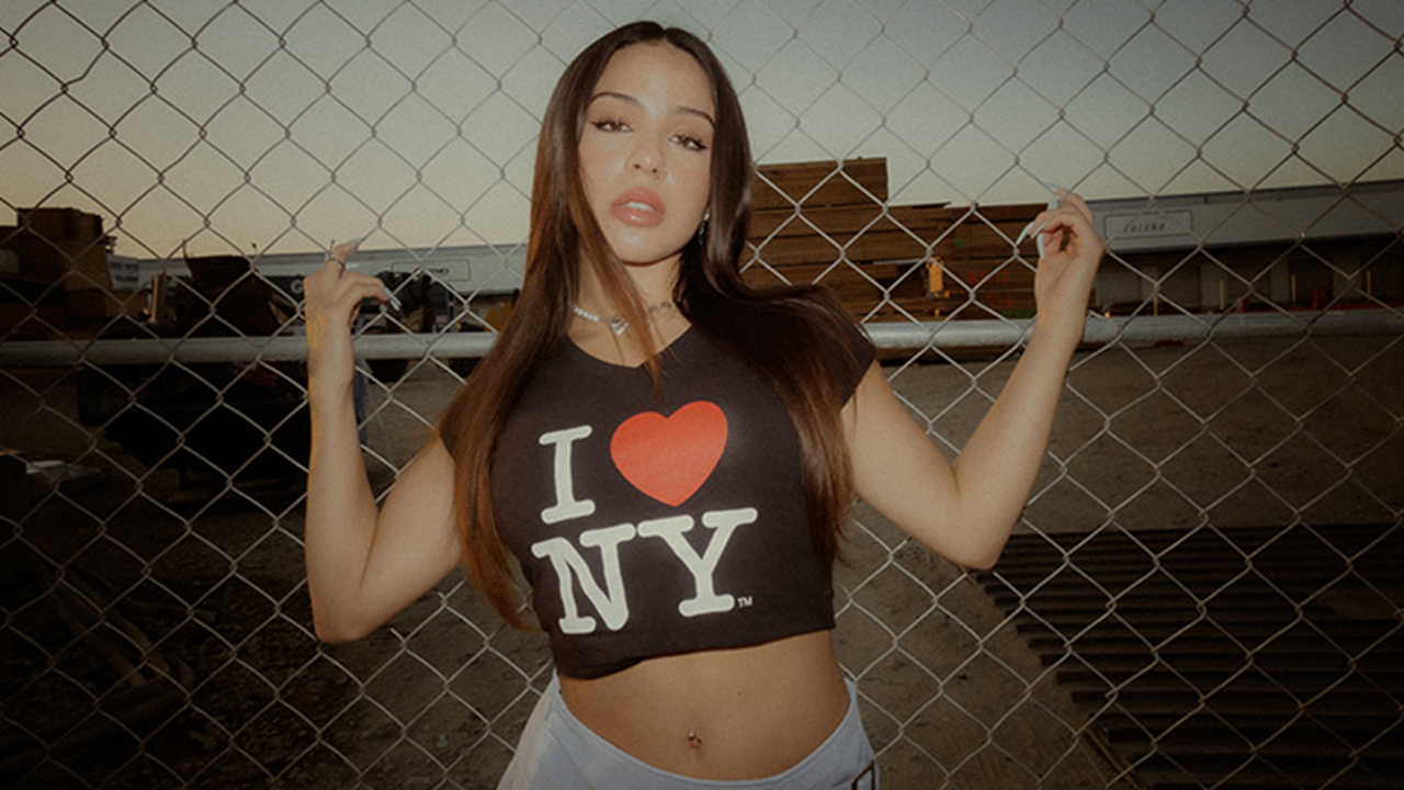 Photo of Jazzy Carrillo leaning on chain link fence behind her in "I love NY" crop top