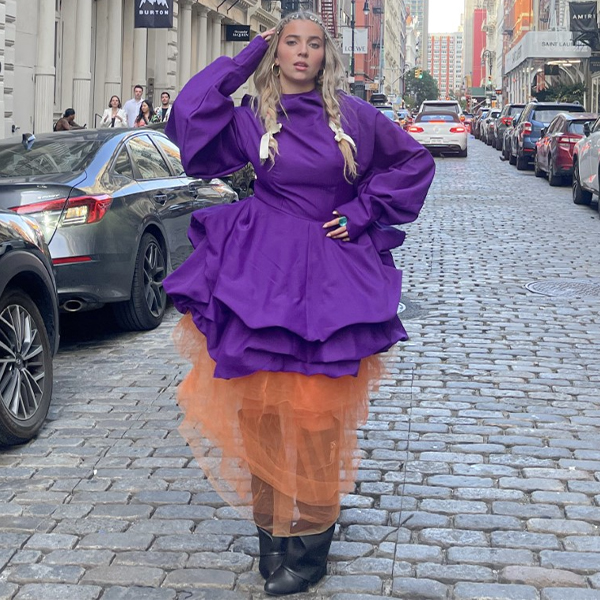 Lexy is on a city street wearing a purple long sleeved dress with an orange tulle underskirt and black boots. She is blonde and has one hand on her hip and the other at her head.