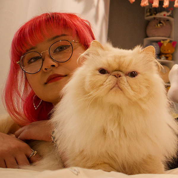 Monica, with pink hair, lays on a bed with her cat held in front of her. Her ankles are crossed behind her.