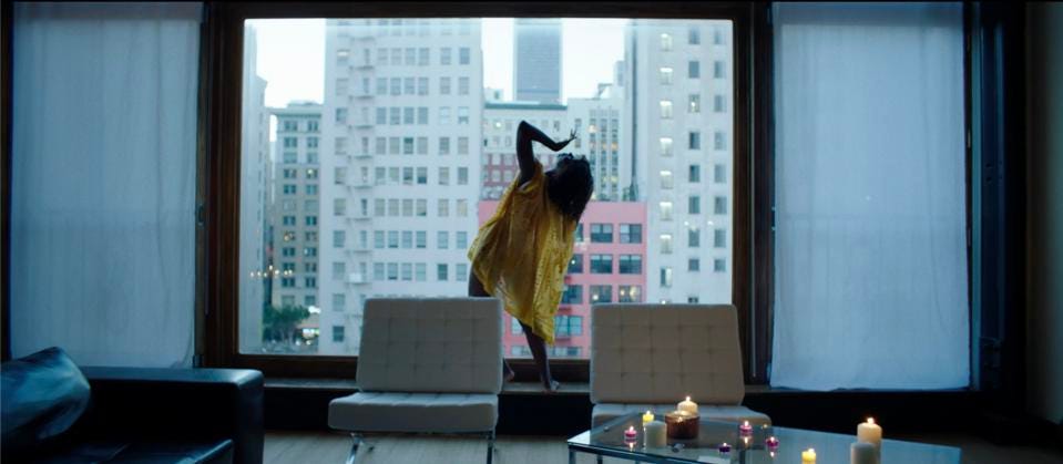 A film still of Natasha Ofili in a yellow dress in front of a large window with a city view