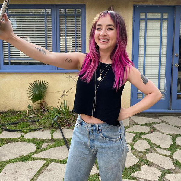 Photo of Cassandra in front of yellow and blue house, taking a selfie but looking at the camera. She has black and pink hair and is wearing a black sleeveless top and jeans.
