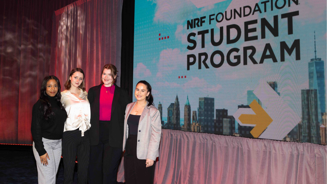 FIDM Students Kiera Berry, Avery Van der Linden, Kristin Hall, and Shannon Curley stand together indoors with a sign behind them that read NRF Student Challenge