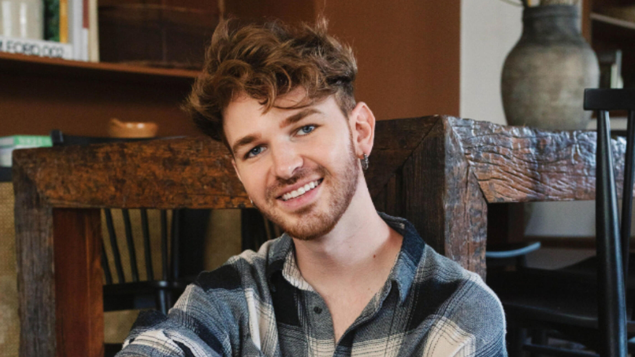 Photo of Drew in plaid shirt leaning on table. His head is cocked to the side, and he has a smile on his face. 