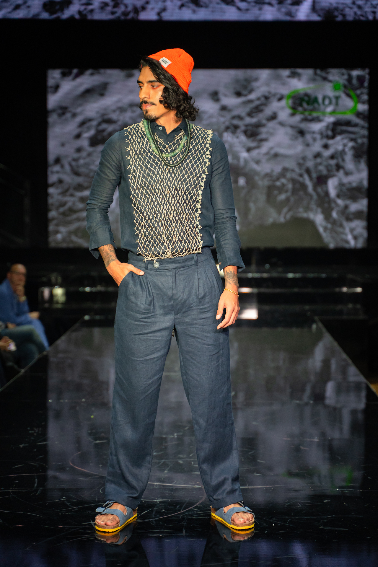 A menswear model stands on the runway in a navy shirt with a net overlay and navy pants designed by Marina Leight
