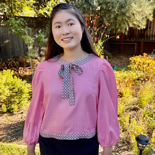 Photo of Isabelle smiling into camera while standing outdoors in front of plants. She is wearing a pink top with a black and white bow at the neckline.