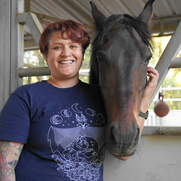 Jessica Mirazo smiles and wears purple t-shirt as she touches the face of a horse outdoors in a stable