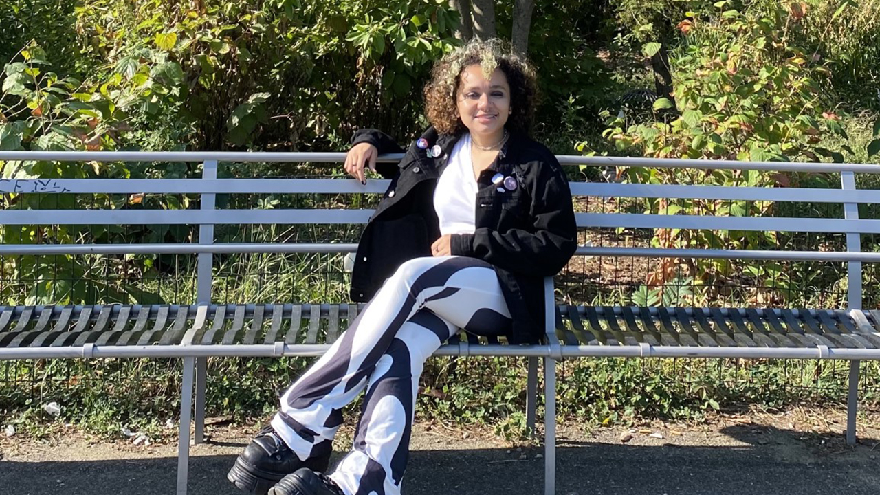 Photo of AJ in black and white pants and black jacket sitting on a park bench outdoors