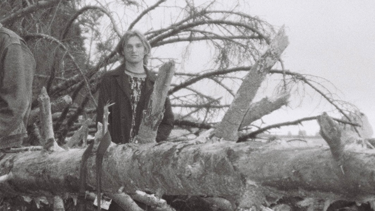 Black and white photo of Evan standing behind a fallen tree outdoors. 