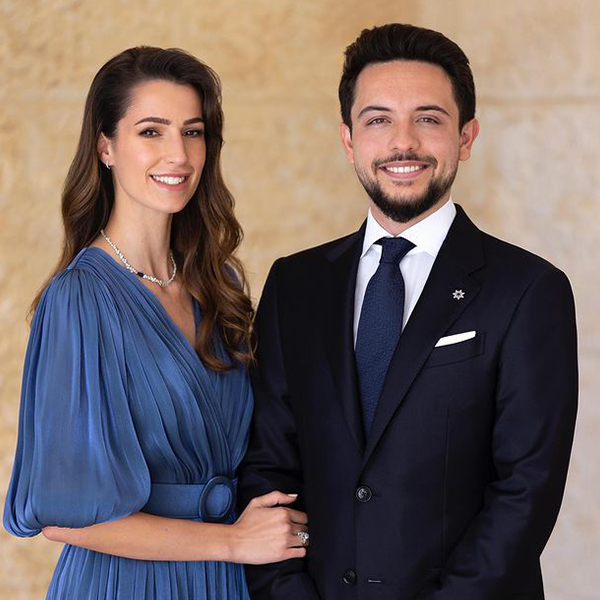 Photo of Rajwa and Hussein in front of beige background. She wears a blue dress and has her hand on his arm. He's in a suit. Both have brown hair.