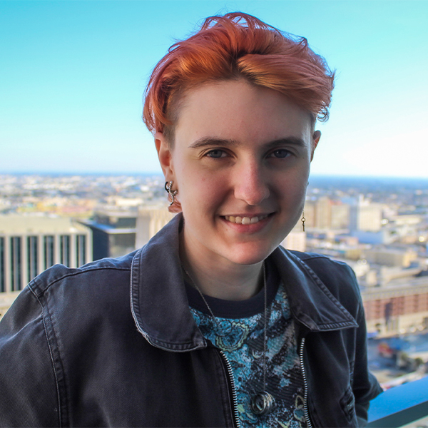 Steven stands on a patio overlooking the city skyline. He has short pink hair and is wearing a denim jacket. He smiles as he looks into the camera.