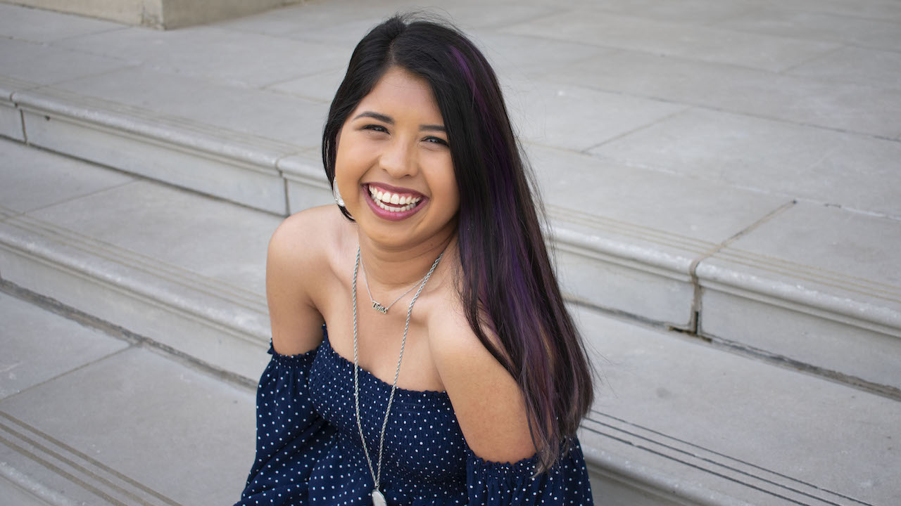 A seated portrait of FIDM Grad Alex Candelas smiling wearing an off the shoulder navy top