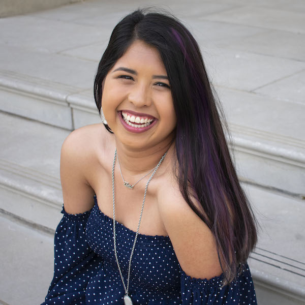 A seated portrait of FIDM Grad Alex Candelas smiling wearing an off the shoulder navy top
