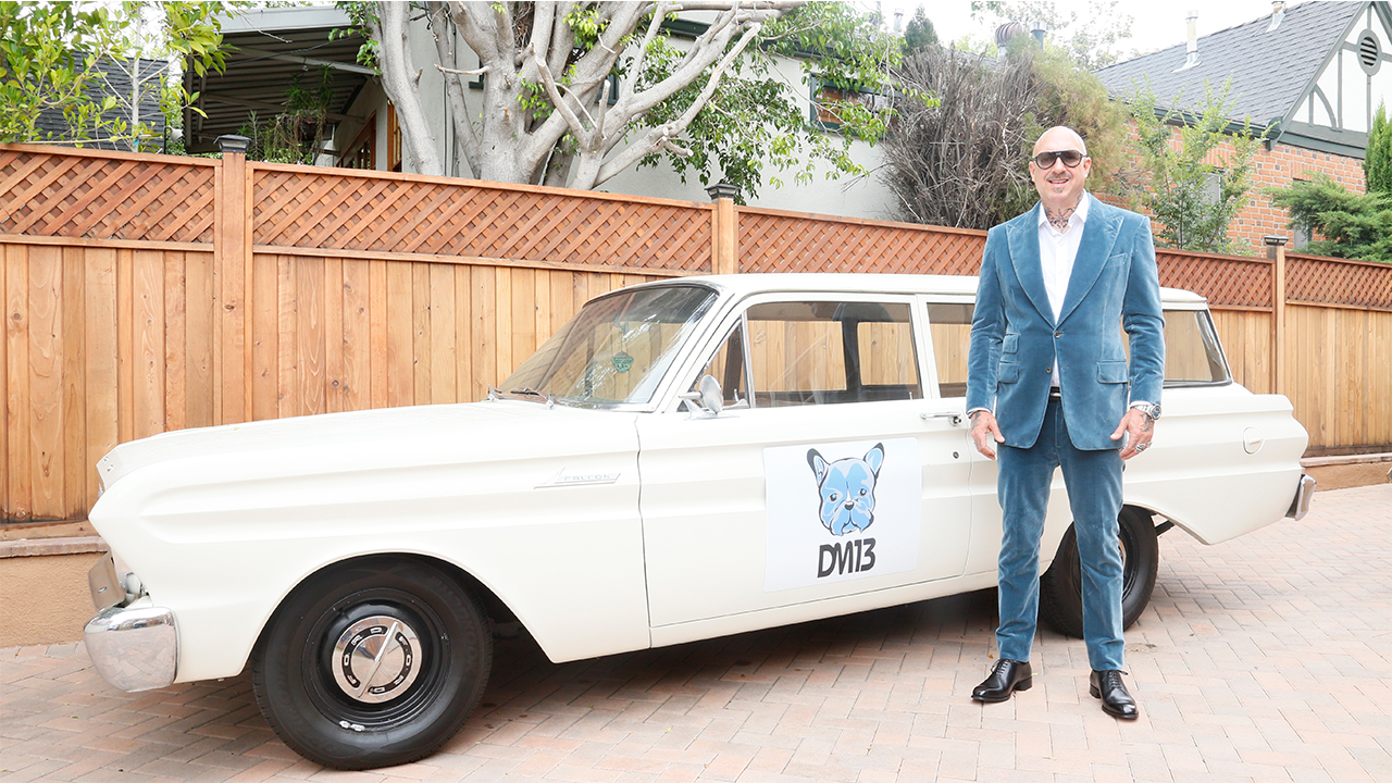 David stands in a blue velvet suit in front of a white car with his company's logo on the door.