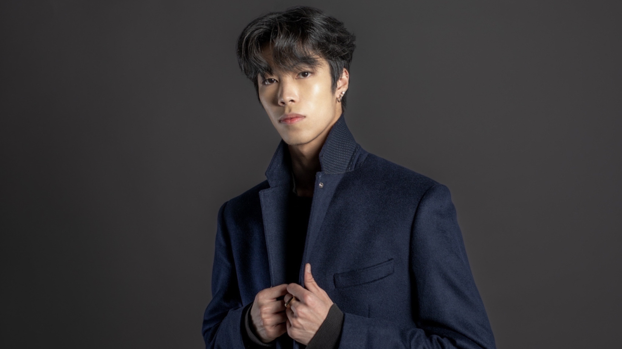 FIDM Advanced Fashion Design Student Max Tran faces the camera indoors against a dark grey wall wearing a navy blue blazer with his hand grasping the collar