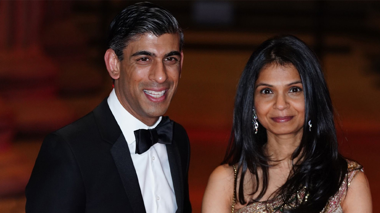 Rishi and Akshata are side by side at an event. He's in a tuxedo, and she's in a champagne colored beaded dress with her dark hair over her shoulders.