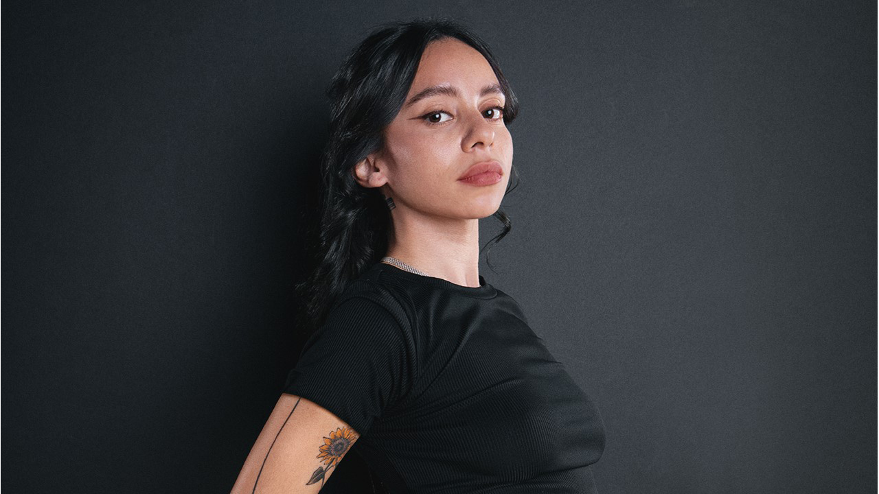 Photo of Gabriela facing right, but looking toward the camera. She wears a black t shirt and has a flower tattoo visible on her arm. She has dark hair and a confident expression on her face. 