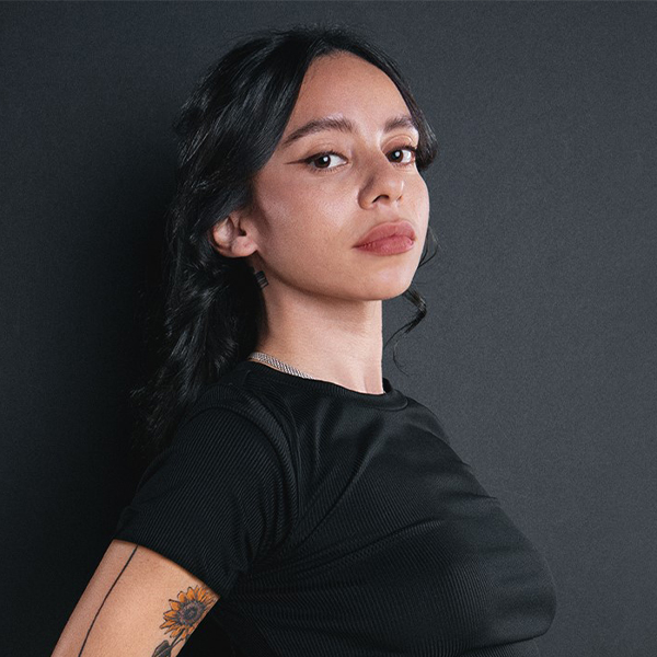 Photo of Gabriela facing right, but looking toward the camera. She wears a black t shirt and has a flower tattoo visible on her arm. She has dark hair and a confident expression on her face. 