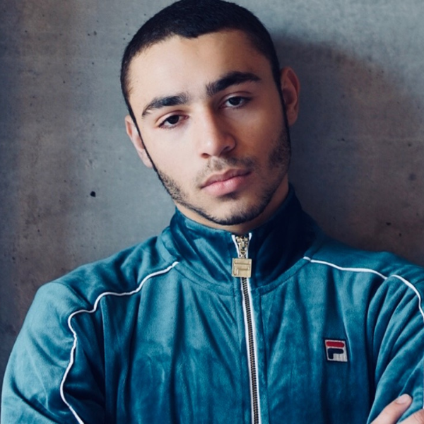 close up of Keagan Roberson standing against a wall with his arms folded across his chest and his head tilted to one side as he wears a zip up blue Fila track jacket