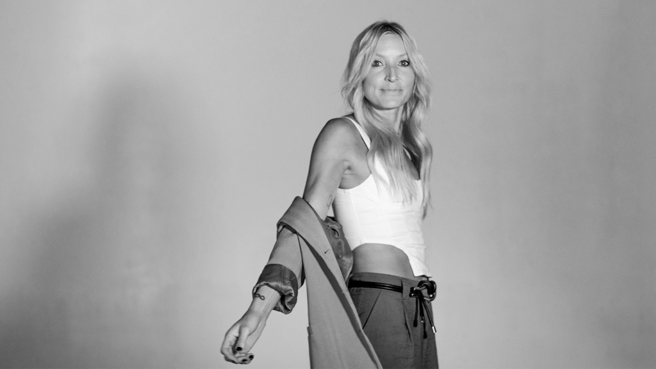 Black and white image of Melissa in front of backdrop, in white tank and trousers, with her jacket sliding down her arms. She is turning to face the camera.