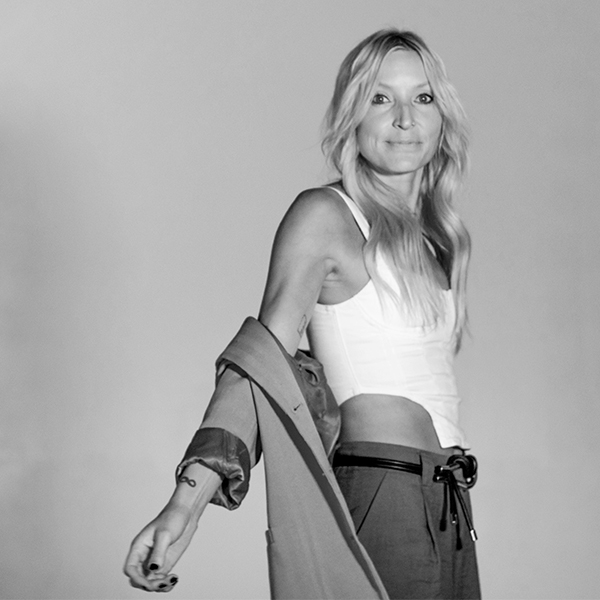 Black and white image of Melissa in front of backdrop, in white tank and trousers, with her jacket sliding down her arms. She is turning to face the camera.