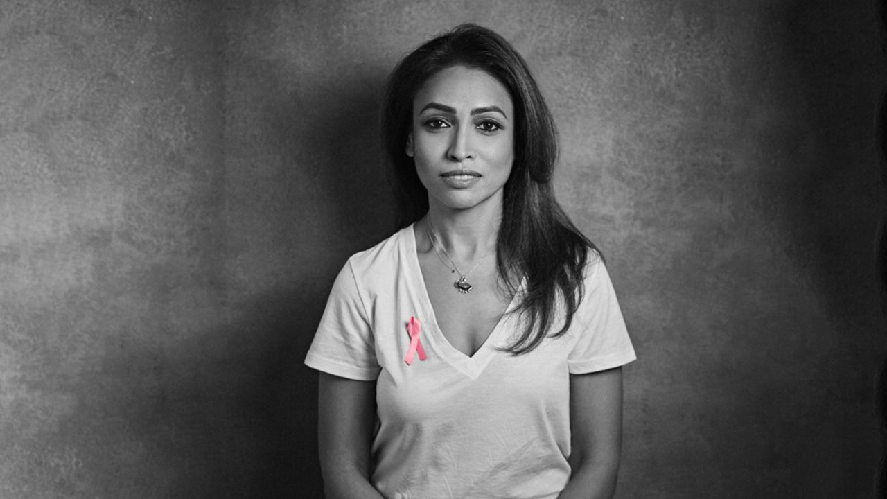 Black and white photo of Surily in white t shirt in front of grey background with pink breast cancer awareness ribbon pinned to her shirt. She has long brown hair over one shoulder.