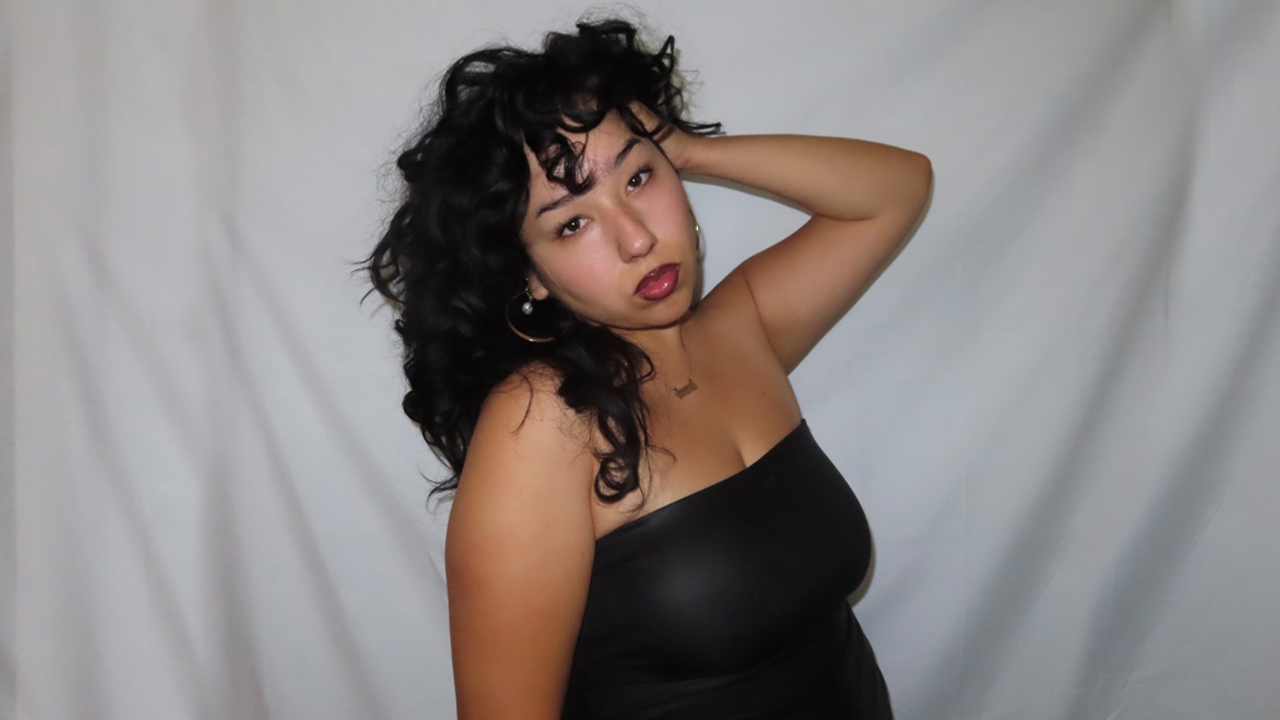 Photo of Alondra in black strapless top and curly hair with right hand at the side of her head looking into camera in front of grey drape