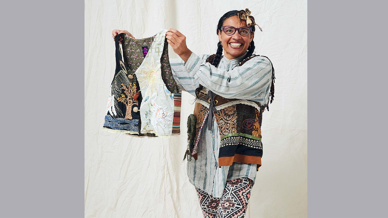 Orquidia wears a chambray shirt, patterned leggings, and a handmade vest and holds up another handmade vest. Both are works of her art.