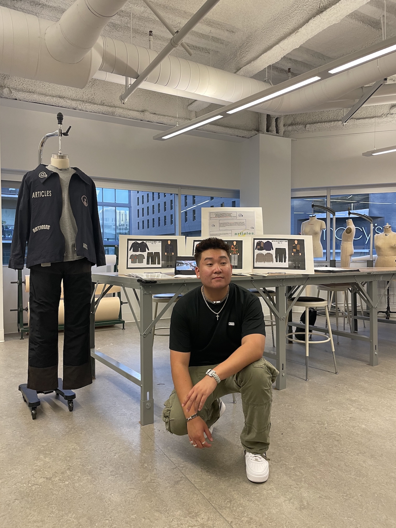FIDM Grad Colin Huang poses with one knee on the ground in front of his menswear designs