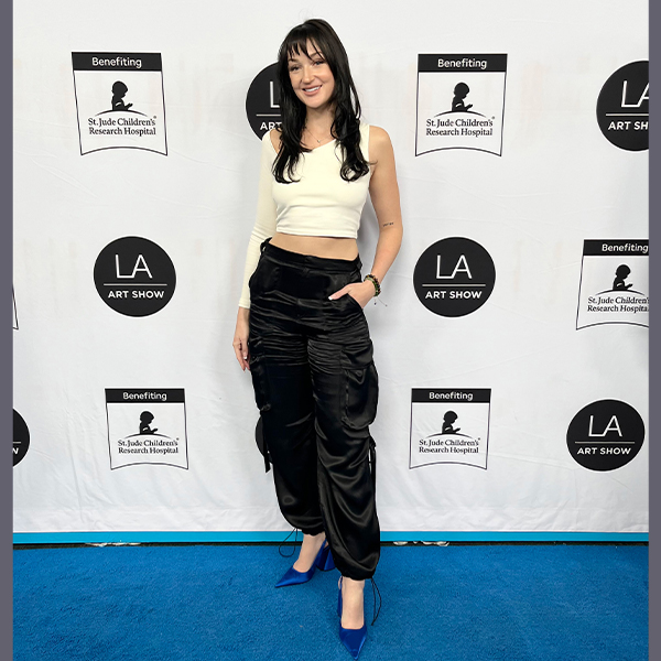 Jordyn is on the LA Art Show Step-and-repeat wearing black satin pants, blue heels, and a white cropped one-sleeve top.. 