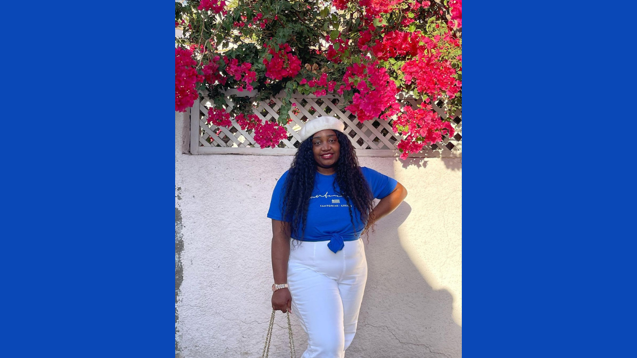 Mazarine Ebengho stands outside in front of bougainvillea wearing a blue tshirt and white pants 